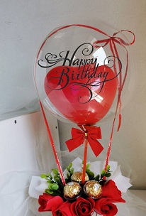 Red heart balloon stuffed inside a clear printed balloon with happy birthday tied with red ribbon to a basket with 3 ferrero chocolates and 5 red roses