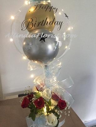 Happy Birthday printed text on the transparent balloon with golden and silver balloons stuffed inside in a box of gold and red net and 6 red roses finally decorated with string lights