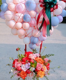 Pink and blue balloons with big ribbon bows 25 in number on the sticks of a basket filled with orange and red flowers