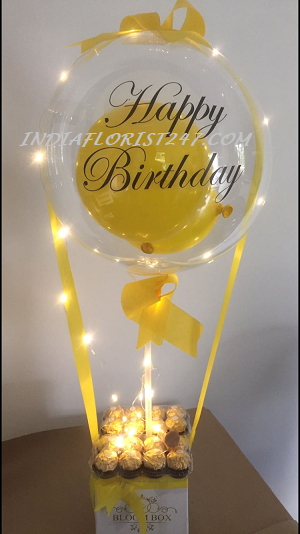 Transparent Happy Birthday with printed message text on balloon stuffed with yellow balloon yellow ribbon with 16 ferrero rocher chocolates and roses finally decorated with LED lights